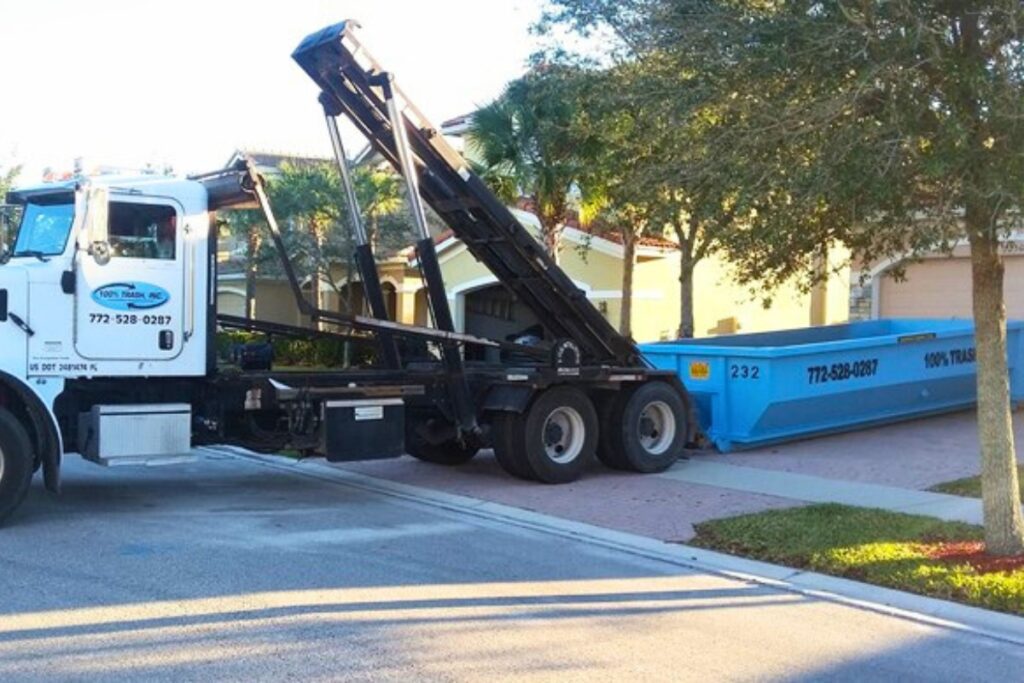 Roll-off truck delivering large blue dumpster on driveway.