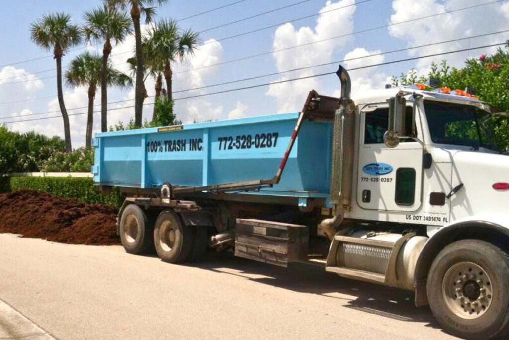 Truck unloading soil on roadside near palm trees.