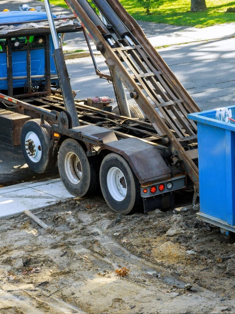 Truck placing blue dumpster on construction site soil.